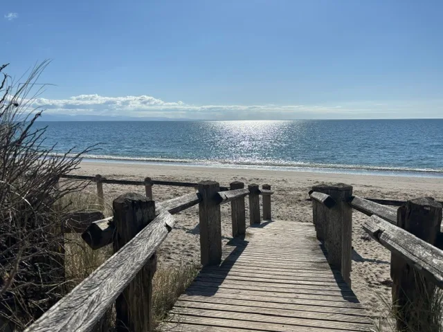A wooden boardwalk leading down onto a sandy beach, with sunlight sparkling on a blue sea