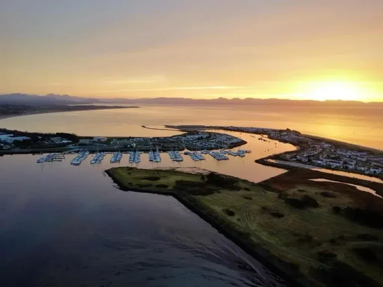 Aerial view at sunset over a harbour full of moored boats, a green spit of land, the town beside it and hills on the horizon