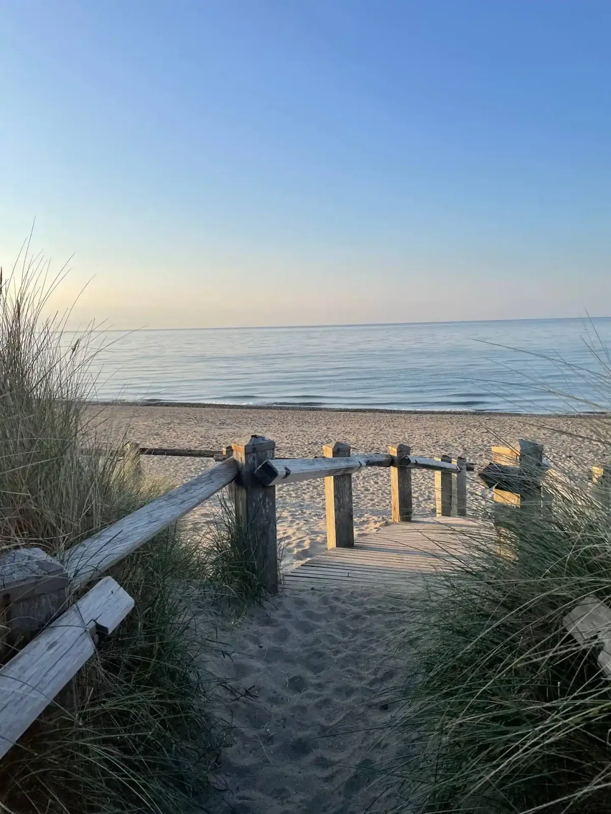 A sandy path between marram grass leading to a weathered boardwalk and a calm sea